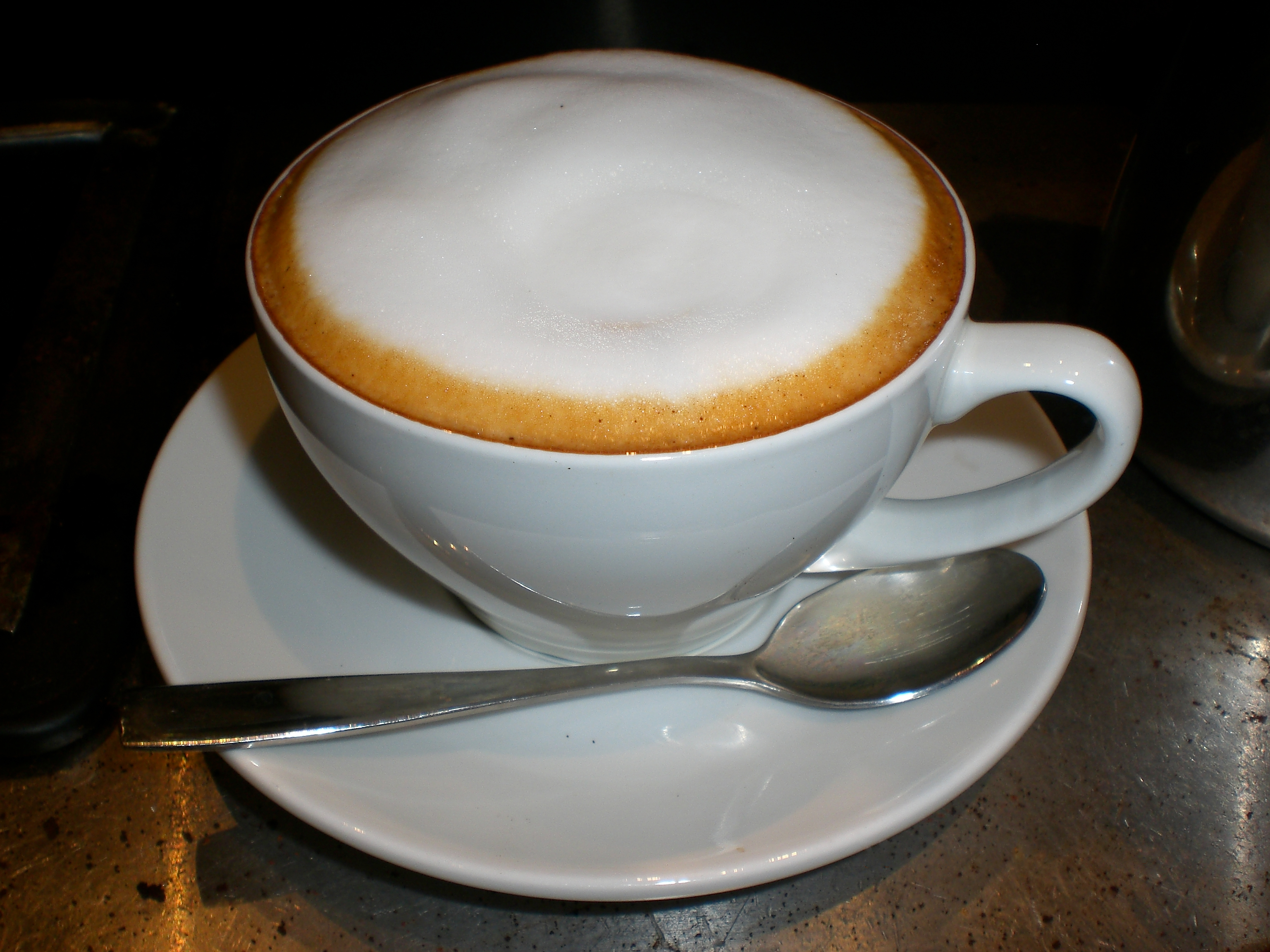 A cup of coffee is shown; a cappuccino. There is a spoon on the plate, and the foam is visible above the side of the cup.